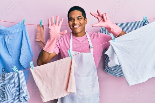 Young handsome hispanic man wearing cleaner apron holding clothes on clothesline showing and pointing up with fingers number eight while smiling confident and happy.