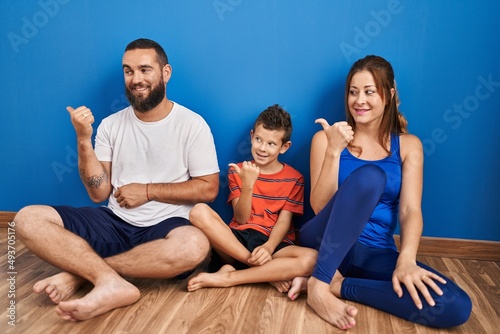 Family of three sitting on the floor at home smiling with happy face looking and pointing to the side with thumb up.