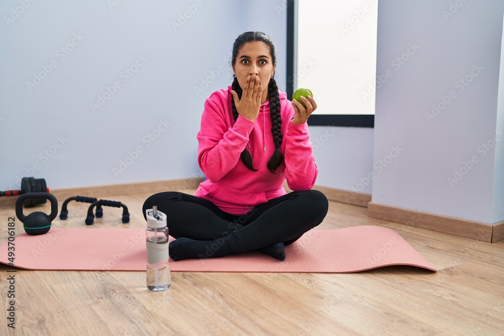 Young hispanic woman sitting on yoga mat eating apple covering mouth with hand, shocked and afraid for mistake. surprised expression Young hispanic woman sitting on yoga mat eating apple covering mouth with hand, shocked and afraid for mistake. surprised expression