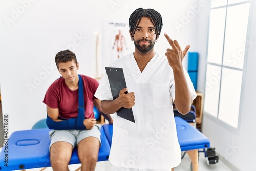 Young hispanic man working at pain recovery clinic with a man with broken arm showing middle finger, impolite and rude fuck off expression