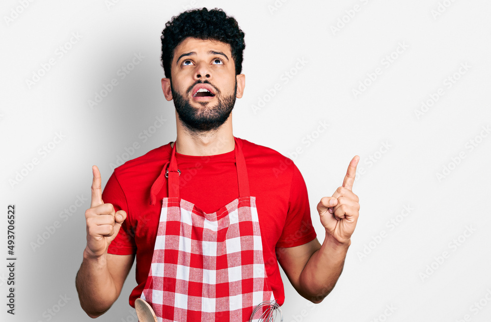 Young arab man with beard wearing cook apron amazed and surprised looking up and pointing with fingers and raised arms. Young arab man with beard wearing cook apron amazed and surprised looking up and pointing with fingers and raised arms.