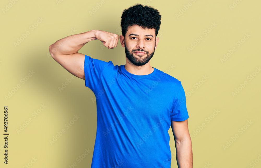 Young arab man with beard wearing casual blue t shirt strong person showing arm muscle, confident and proud of power Young arab man with beard wearing casual blue t shirt strong person showing arm muscle, confident and proud of power