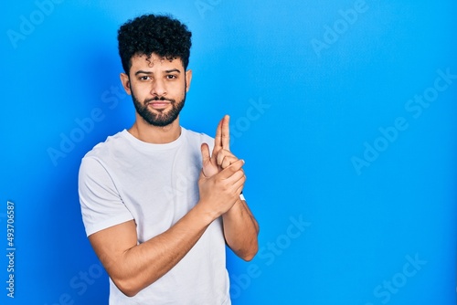 Young arab man with beard wearing casual white t shirt holding symbolic gun with hand gesture, playing killing shooting weapons, angry face