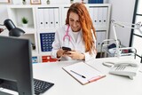 Young redhead woman wearing doctor uniform using smartphone at hospital