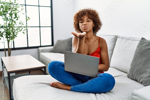 Young african american woman sitting on the sofa at home using laptop looking at the camera blowing a kiss with hand on air being lovely and sexy. love expression.