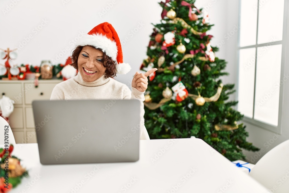 Middle age hispanic woman using laptop sitting by christmas tree smiling happy pointing with hand and finger to the side Middle age hispanic woman using laptop sitting by christmas tree smiling happy pointing with hand and finger to the side