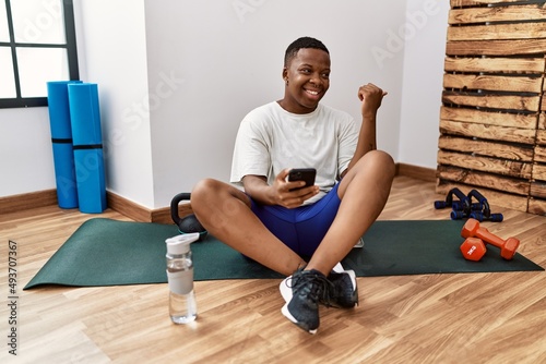 Young african man sitting on training mat at the gym using smartphone smiling with happy face looking and pointing to the side with thumb up.