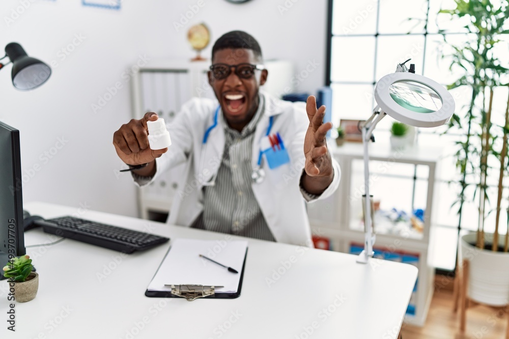 Young african american doctor man wearing doctor uniform holding prescription pills at the clinic celebrating victory with happy smile and winner expression with raised hands