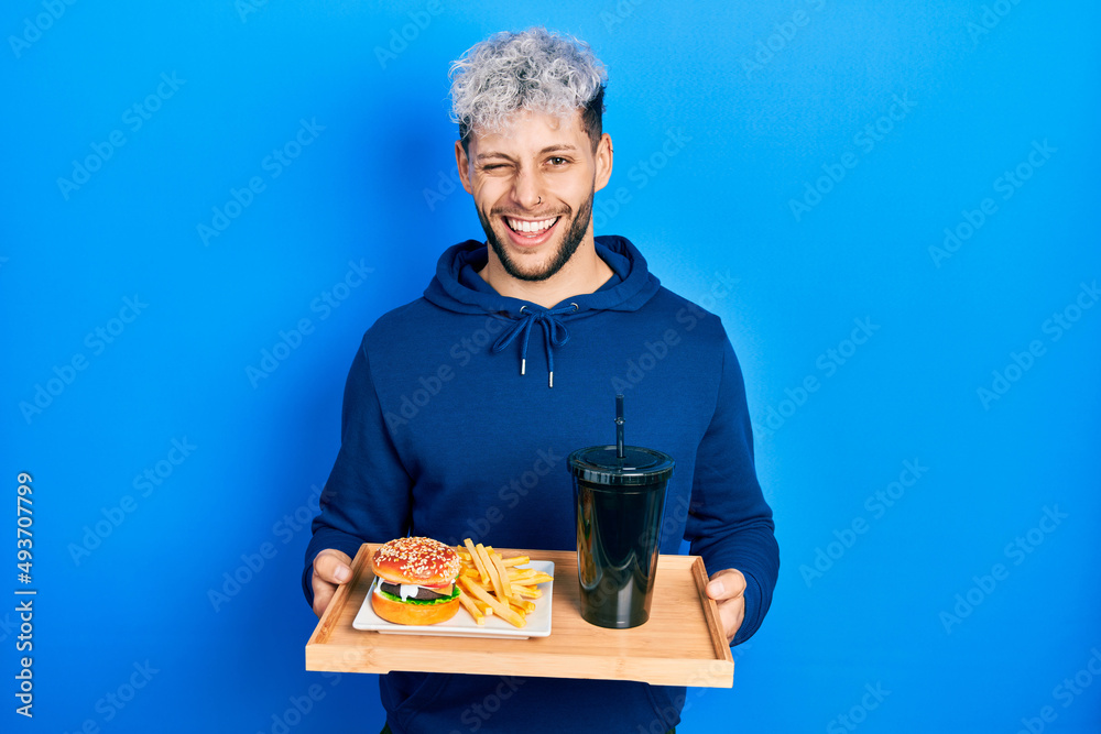 Young hispanic man with modern dyed hair eating a tasty classic burger with fries and soda winking looking at the camera with sexy expression, cheerful and happy face. Young hispanic man with modern dyed hair eating a tasty classic burger with fries and soda winking looking at the camera with sexy expression, cheerful and happy face.