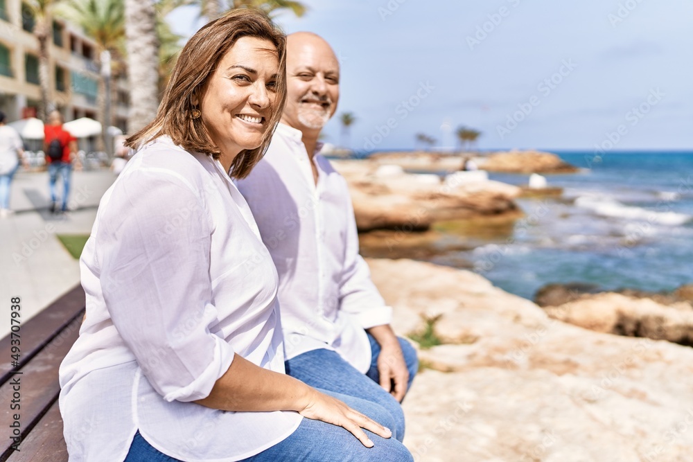 Middle age hispanic couple of husband and wife together sitting by the beach on a sunny day. Hugging in love on vacation to the seaside.