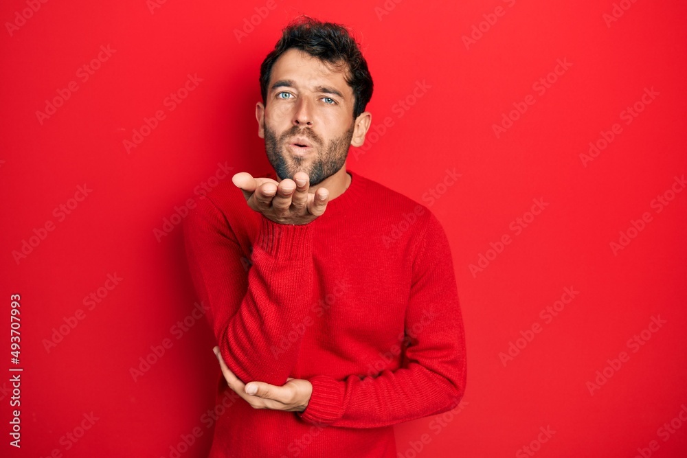 Handsome man with beard wearing casual red sweater looking at the camera blowing a kiss with hand on air being lovely and sexy. love expression. Handsome man with beard wearing casual red sweater looking at the camera blowing a kiss with hand on air being lovely and sexy. love expression.