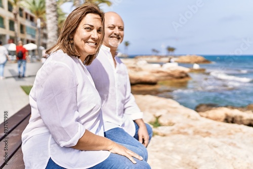 Middle age hispanic couple of husband and wife together sitting by the beach on a sunny day. Hugging in love on vacation to the seaside.