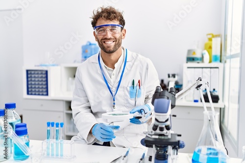 Young hispanic man wearing scientist uniform using loupe at laboratory