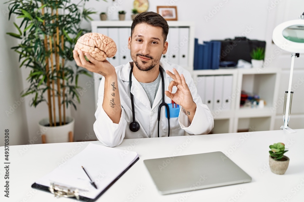 Young doctor holding brain at medical clinic doing ok sign with fingers, smiling friendly gesturing excellent symbol