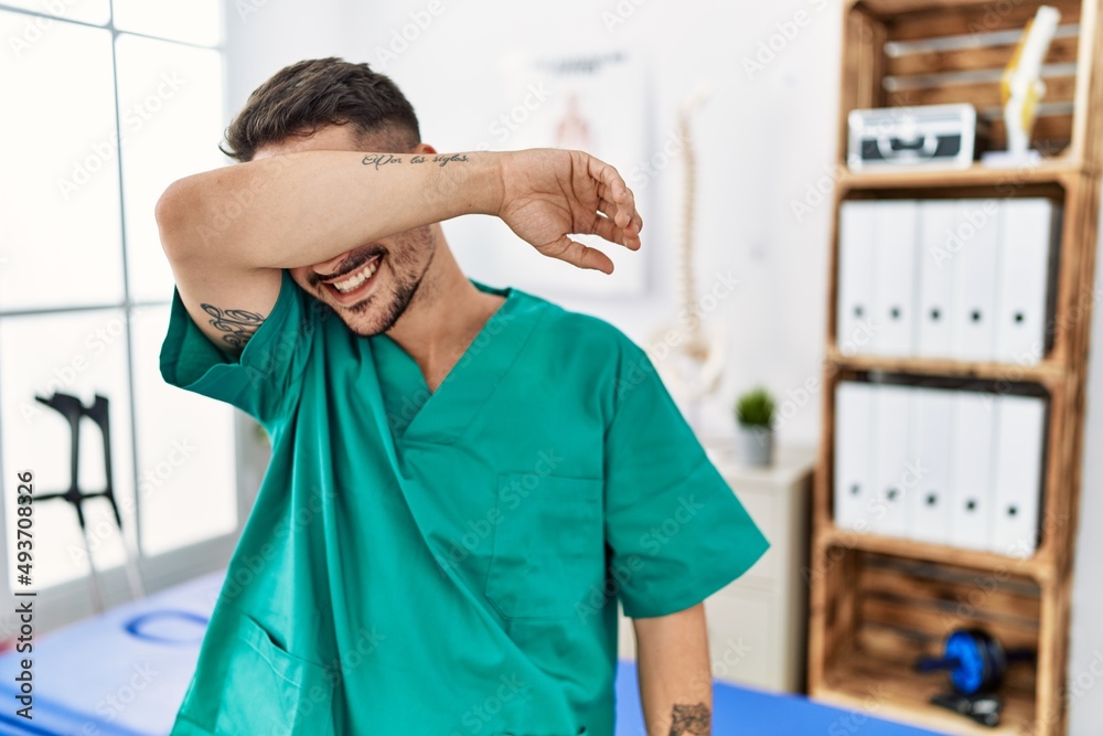 Obraz premium Young physiotherapist man working at pain recovery clinic covering eyes with arm smiling cheerful and funny. blind concept.