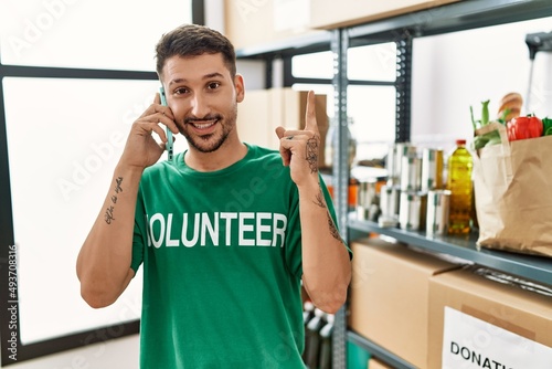 Young hispanic man wearing volunteer t shirt speaking on the phone smiling happy pointing with hand and finger to the side