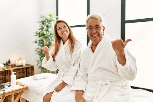 Middle age hispanic couple wearing bathrobe at wellness spa smiling with happy face looking and pointing to the side with thumb up.