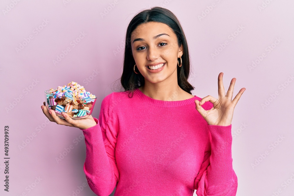 Young hispanic woman holding bowl of sugar candy doing ok sign with fingers, smiling friendly gesturing excellent symbol Young hispanic woman holding bowl of sugar candy doing ok sign with fingers, smiling friendly gesturing excellent symbol