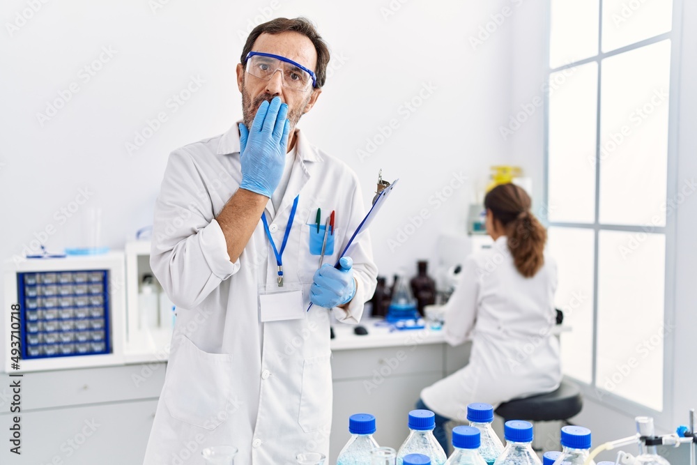 Middle age man working at scientist laboratory covering mouth with hand, shocked and afraid for mistake. surprised expression Middle age man working at scientist laboratory covering mouth with hand, shocked and afraid for mistake. surprised expression