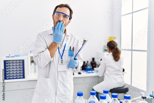 Middle age man working at scientist laboratory covering mouth with hand, shocked and afraid for mistake. surprised expression