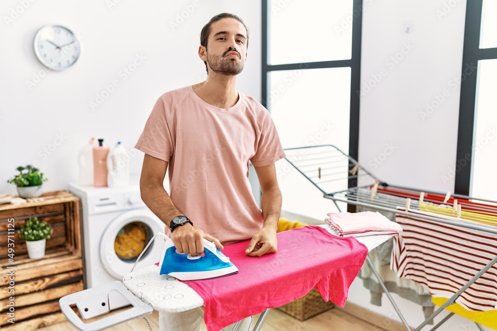 Obraz premium Young hispanic man ironing clothes at home looking at the camera blowing a kiss on air being lovely and sexy. love expression.