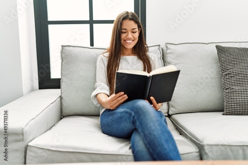 Young latin woman reading book sitting on sofa at home