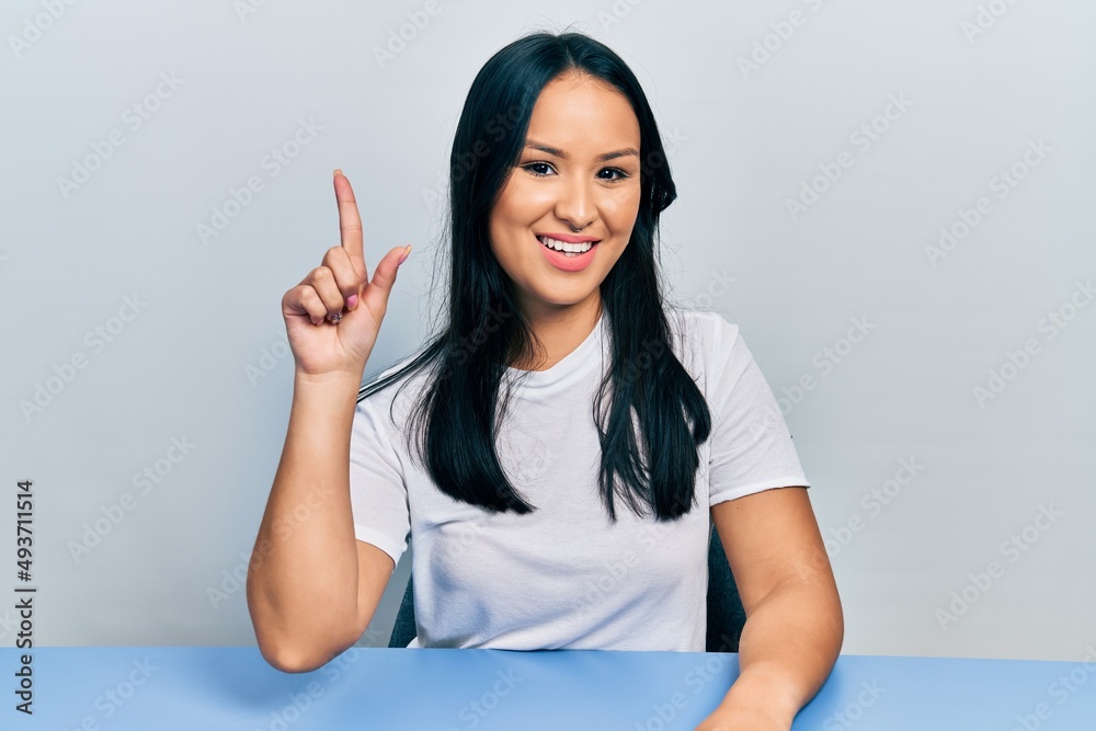 Beautiful hispanic woman with nose piercing wearing casual white t shirt sitting on the table smiling with an idea or question pointing finger up with happy face, number one Beautiful hispanic woman with nose piercing wearing casual white t shirt sitting on the table smiling with an idea or question pointing finger up with happy face, number one