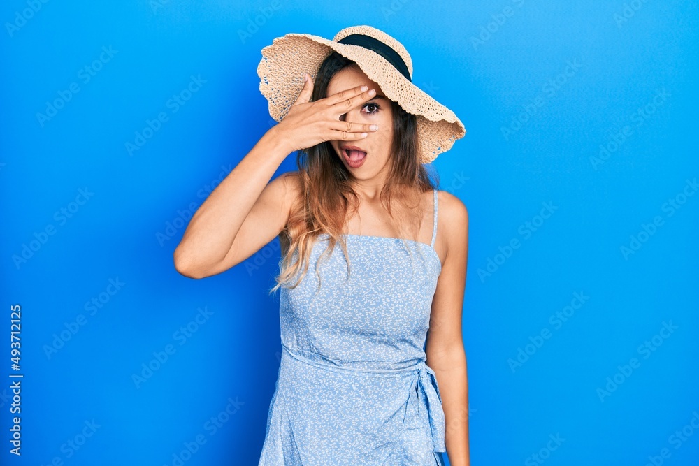 Young hispanic girl wearing summer hat peeking in shock covering face and eyes with hand, looking through fingers with embarrassed expression. Young hispanic girl wearing summer hat peeking in shock covering face and eyes with hand, looking through fingers with embarrassed expression.