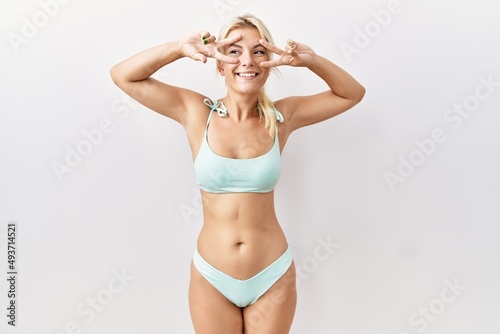 Young caucasian woman wearing bikini over isolated background doing peace symbol with fingers over face, smiling cheerful showing victory