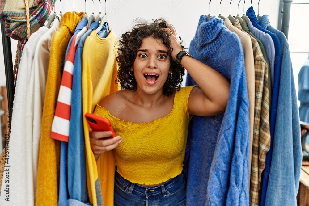 Fototapeta premium Young hispanic woman searching clothes on clothing rack using smartphone crazy and scared with hands on head, afraid and surprised of shock with open mouth