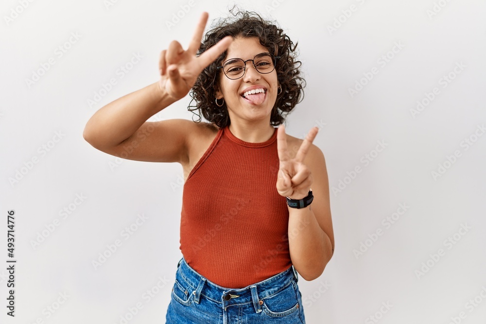 Young hispanic woman wearing glasses standing over isolated background smiling with tongue out showing fingers of both hands doing victory sign. number two. Young hispanic woman wearing glasses standing over isolated background smiling with tongue out showing fingers of both hands doing victory sign. number two.