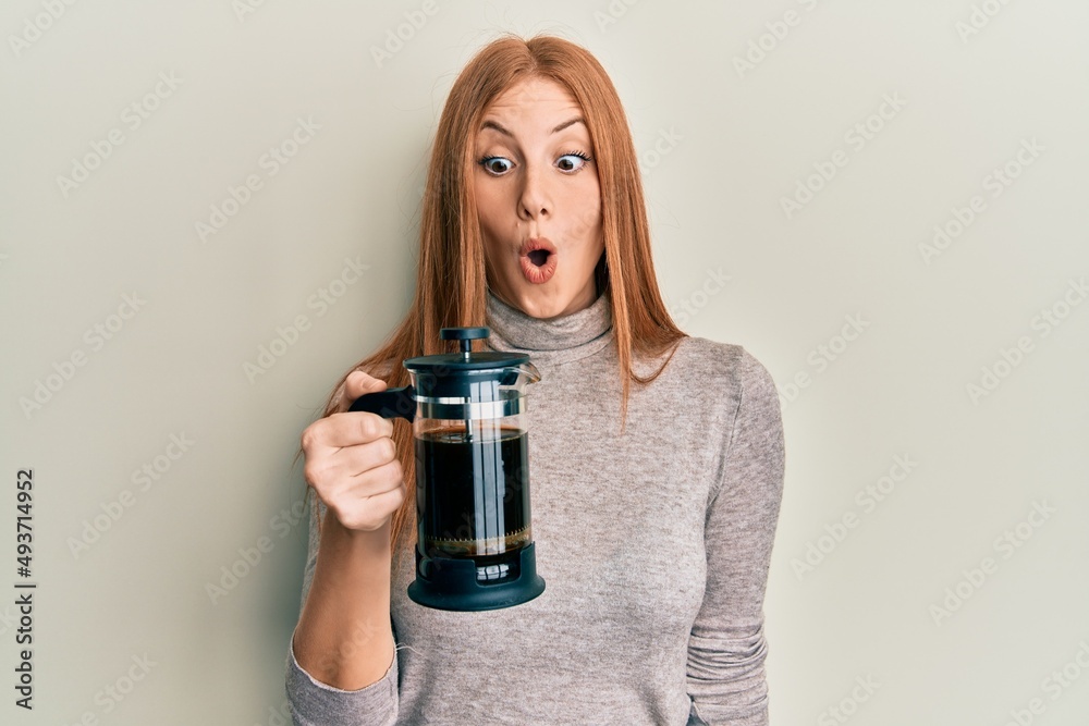Young irish woman holding french coffee maker scared and amazed with open mouth for surprise, disbelief face Young irish woman holding french coffee maker scared and amazed with open mouth for surprise, disbelief face