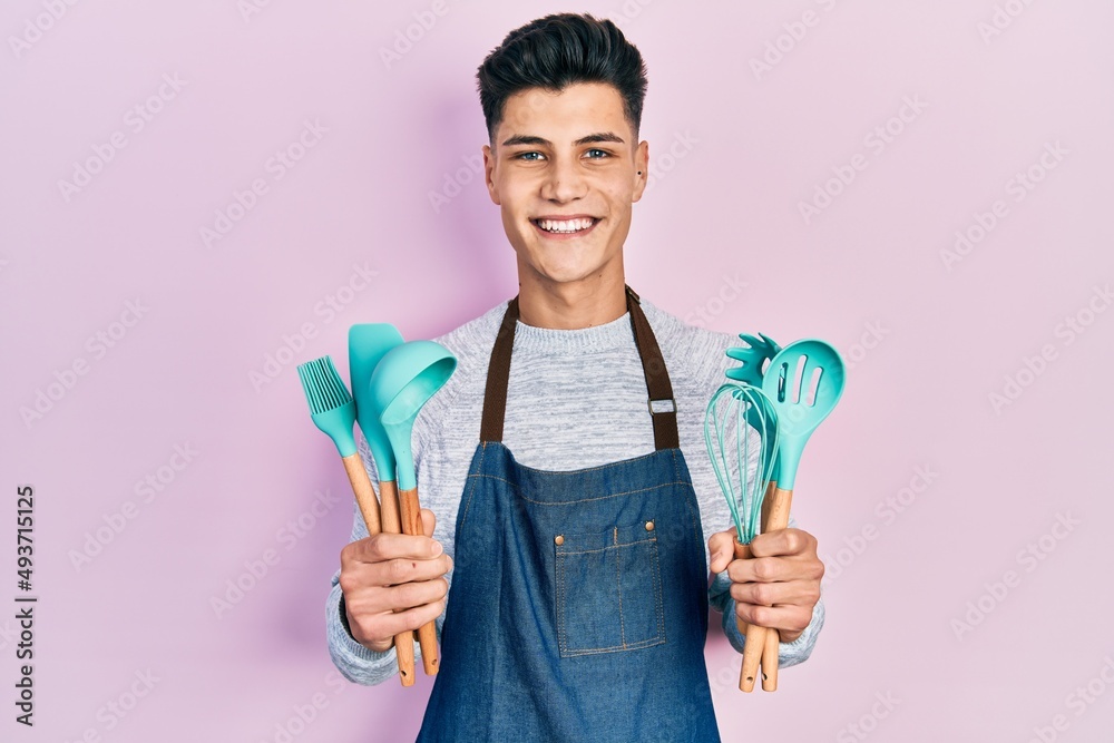 Young hispanic man wearing professional baker apron holding cooking tools smiling with a happy and cool smile on face. showing teeth. Young hispanic man wearing professional baker apron holding cooking tools smiling with a happy and cool smile on face. showing teeth.
