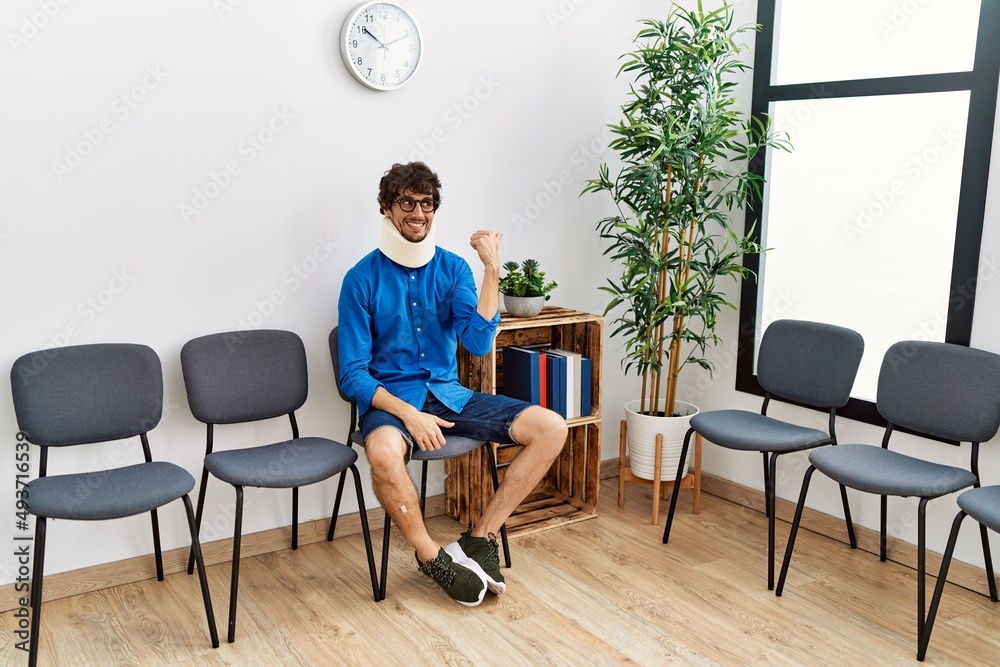 Fototapeta premium Young hispanic man sitting at doctor waiting room with neck injury smiling with happy face looking and pointing to the side with thumb up.