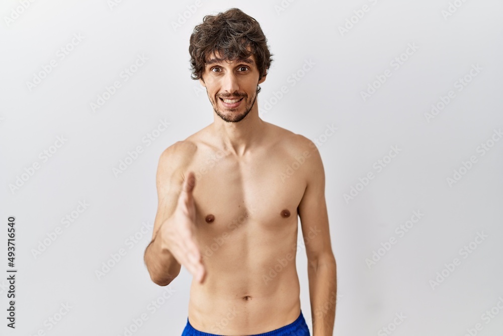 Fototapeta premium Young hispanic man standing shirtless over isolated, background smiling friendly offering handshake as greeting and welcoming. successful business.