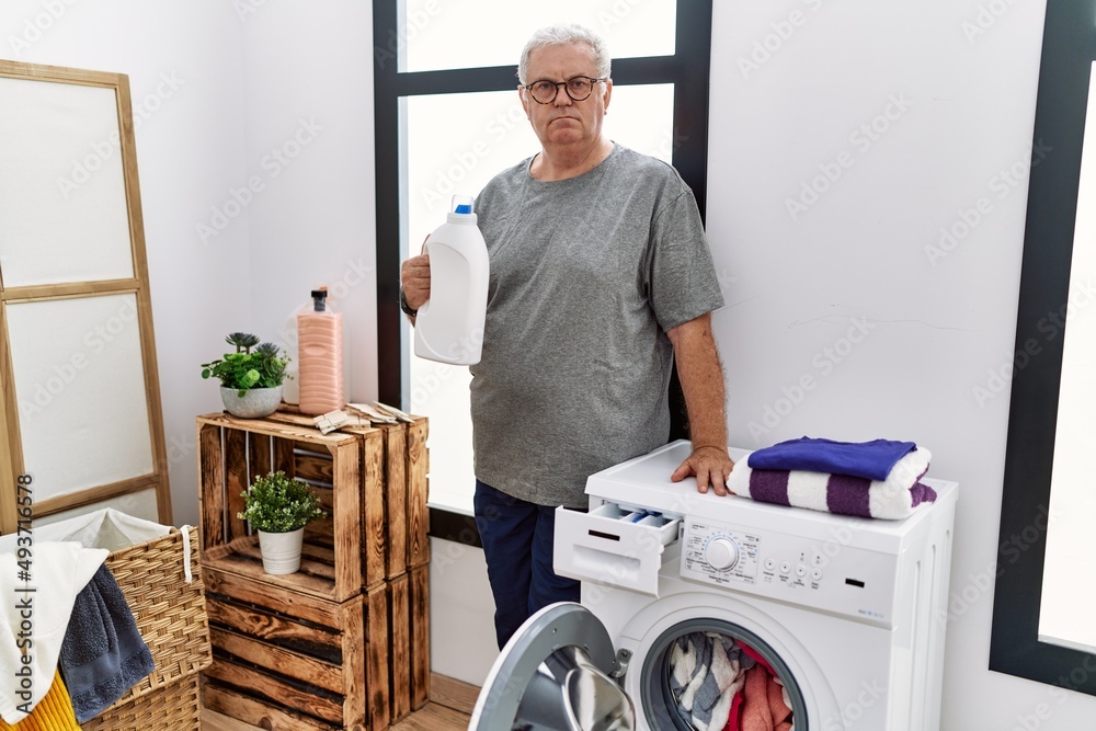 Obraz premium Senior caucasian man doing laundry holding detergent bottle thinking attitude and sober expression looking self confident