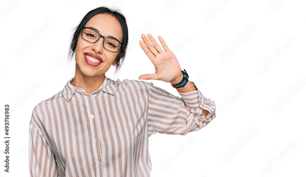 Young hispanic girl wearing casual clothes and glasses waiving saying hello happy and smiling, friendly welcome gesture Young hispanic girl wearing casual clothes and glasses waiving saying hello happy and smiling, friendly welcome gesture