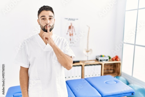 Young handsome man with beard working at pain recovery clinic asking to be quiet with finger on lips. silence and secret concept.