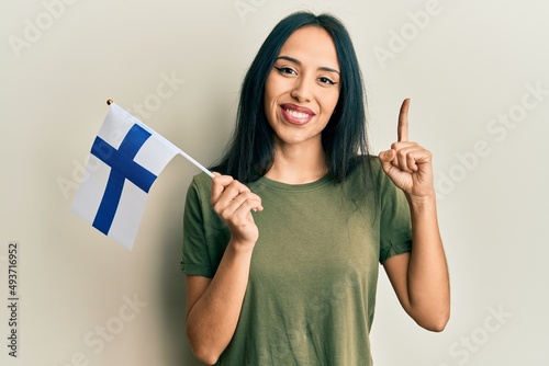 Young hispanic girl holding finland flag smiling with an idea or question pointing finger with happy face, number one