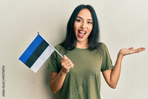 Young hispanic girl holding estonia flag celebrating achievement with happy smile and winner expression with raised hand