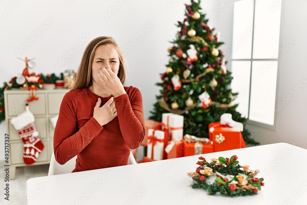 Caucasian young blonde woman sitting on the table by christmas tree smelling something stinky and disgusting, intolerable smell, holding breath with fingers on nose. bad smell Caucasian young blonde woman sitting on the table by christmas tree smelling something stinky and disgusting, intolerable smell, holding breath with fingers on nose. bad smell