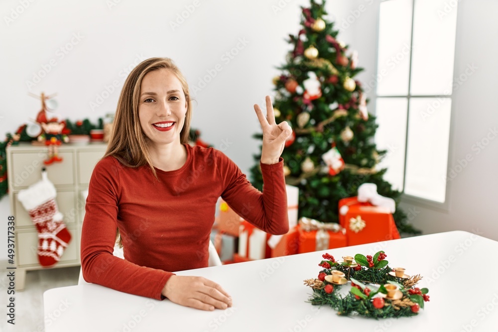 Caucasian young blonde woman sitting on the table by christmas tree showing and pointing up with fingers number two while smiling confident and happy. Caucasian young blonde woman sitting on the table by christmas tree showing and pointing up with fingers number two while smiling confident and happy.