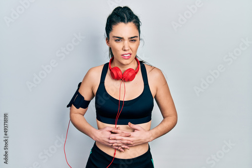 Young hispanic girl wearing gym clothes and using headphones with hand on stomach because indigestion, painful illness feeling unwell. ache concept.