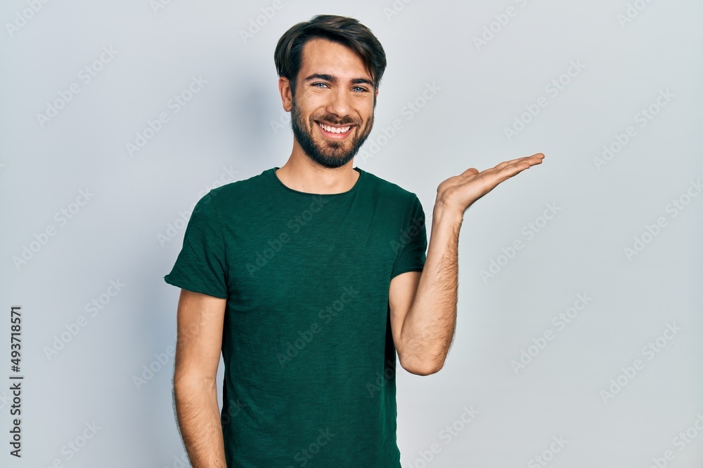 Fototapeta premium Young hispanic man wearing casual white tshirt smiling cheerful presenting and pointing with palm of hand looking at the camera.