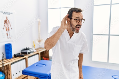 Middle age man with beard working at pain recovery clinic smiling with hand over ear listening an hearing to rumor or gossip. deafness concept.