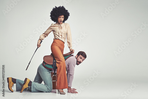 Get moving. A studio shot of an attractive woman in 70s wear riding a handsome man wearing a saddle while using a riding crop.