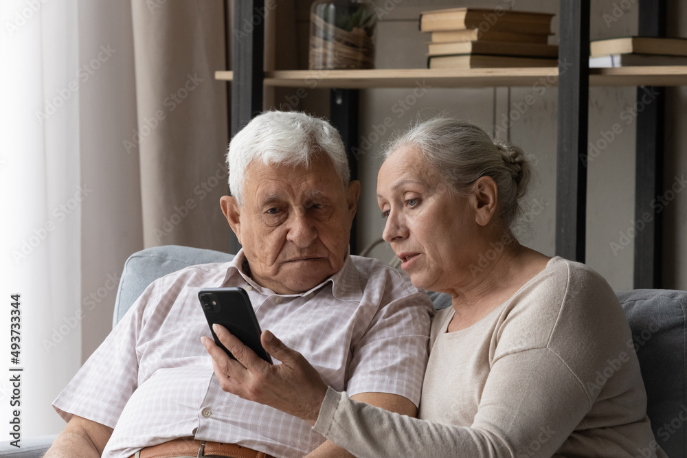 Senior couple discuss mobile app seated on sofa at home, staring at smart phone screen learn new software. Loving wife explains modern device usage to mature husband. Help, tech and older gen concept Senior couple discuss mobile app seated on sofa at home, staring at smart phone screen learn new software. Loving wife explains modern device usage to mature husband. Help, tech and older gen concept
