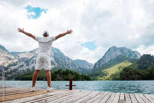 A man in white stands against the backdrop of a lake and mountains with his hands up, a beautiful landscape, rear view. The concept of freedom, happiness, success, victory.