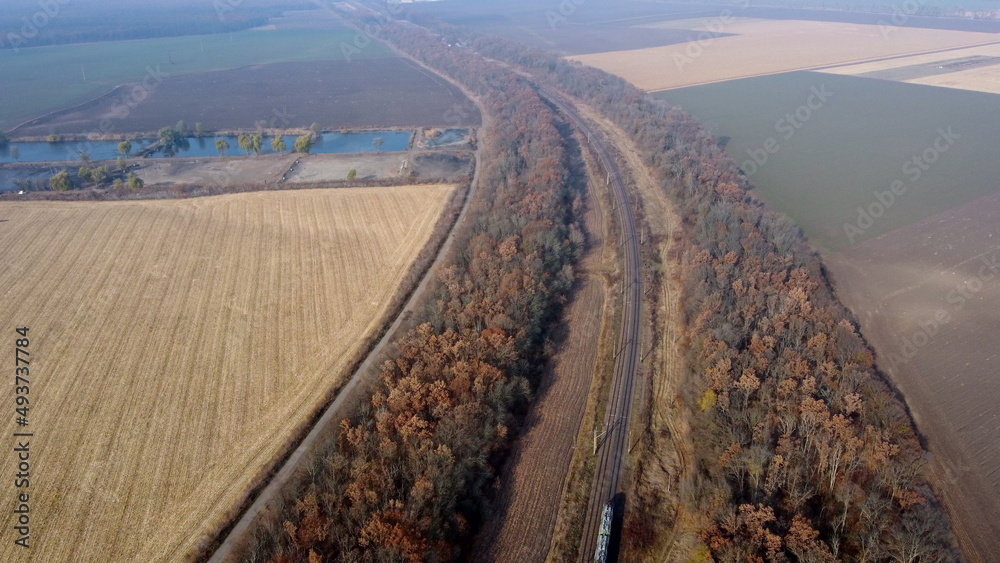 Panoramic View of Moving Freight Train Along Railway Tracks Among Trees Between Agricultural Fields on an Autumn Day. Landscape Freight Cars or Railway Wagon Rides on Railroad. Top View Rail freight