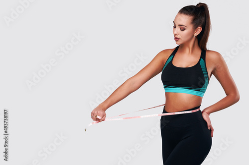 Strong attractive white fitness female in sport suit measuring waist on gray isolated background © khmelev